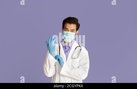 Schutz vor Viruserkrankungen. Indischer Arzt trägt medizinische Maske, Laborkittel und Gummihandschuhe auf violettem Hintergrund Stockfoto