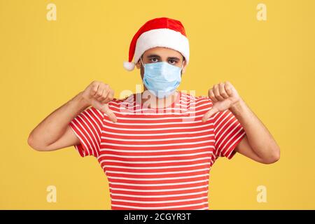 Trauriger junger Mann in weihnachtsmann Hut mit chirurgischen medizinischen Maske zeigt Daumen nach unten Blick auf die Kamera, unzufrieden mit Feiertage Feier in Gesichtsmaske Stockfoto