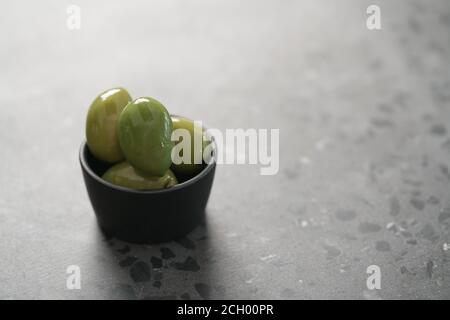 Große grüne Oliven in schwarzer Schale auf Beton Hintergrund, flacher Fokus Stockfoto