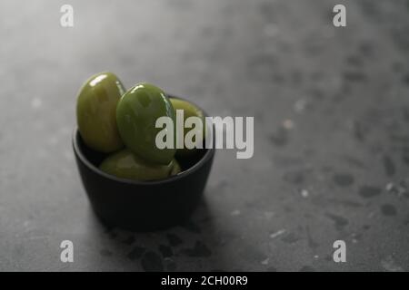 Große grüne Oliven in schwarzer Schale auf Beton Hintergrund, flacher Fokus Stockfoto