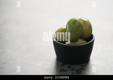Große grüne Oliven in schwarzer Schale auf Beton Hintergrund, flacher Fokus Stockfoto