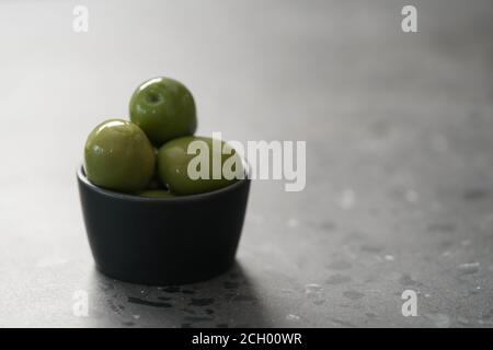 Große grüne Oliven in schwarzer Schale auf Beton Hintergrund, flacher Fokus Stockfoto
