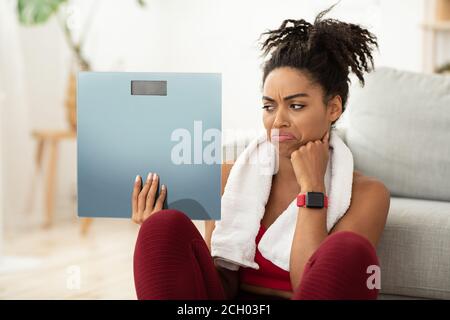 Unzufrieden African Girl Holding Gewichtswaagen Sitzen Auf Dem Boden Zu Hause Stockfoto