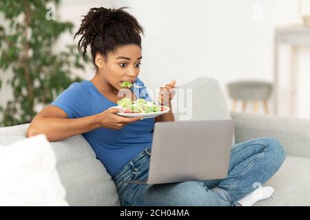 Schockiert Schwarze Frau Essen Gemüse Salat Sitzen Mit Laptop Indoor Stockfoto