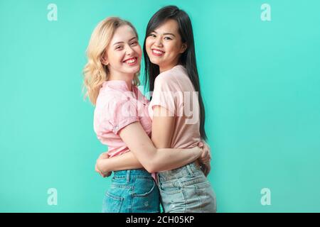 Zwei junge multikulturelle Freundinnen umarmen einander isoliert auf blauem Studiohintergrund. Glücklich kaukasischen und asiatischen Frauen umarmt, Feier, Stockfoto