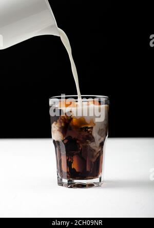 Gießen Sie frische Milch in ein Glas schwarzen Kaffee mit Eiswürfeln auf weißem Brett und schwarzem Hintergrund. Stockfoto