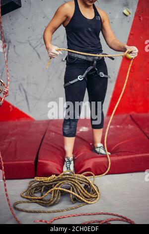 Portrait der unrekonizierbaren Kletterin im Sicherheitsgeschirr gegen Kletterwand, nach dem Training im Kletterverein Sicherheitsleine binden, beschnittene Ansicht Stockfoto