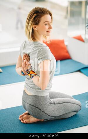 Junge Frau macht Yoga-Übung auf Matte in Virasana oder Held Pose mit umgekehrtem Gebet oder namaste im Studio vor einem melierten Hintergrund von hinten. Stockfoto