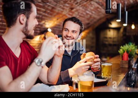 Zwei glückliche männliche Freunde, die in der beliebten Burger Bar zu Abend essen. Junge Leute sitzen, reden und essen Burger, Bier trinken. Stockfoto