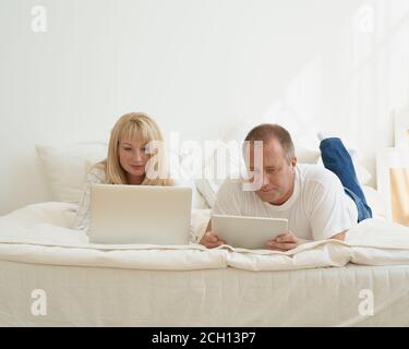 Porträt eines reifen Paares mit digitaler sucht zu Hause. Schöner Mann mit Tablet und attraktive Frau mittleren Alters mit Laptop verbringen Zeit zusammen, während im Bett liegen. Stockfoto