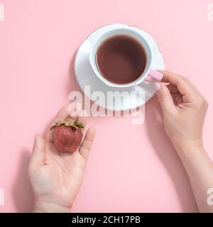 Damenhände mit einer Tasse Tee und Erdbeeren auf einem rosa Hintergrund. Weiße Tasse Tee auf einer weißen Untertasse. Draufsicht Stockfoto