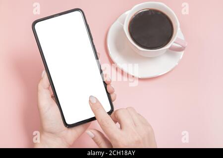 Handy, Smartphone. Die Hände der Frauen halten ein Handy mit einem leeren Bildschirm auf einem rosa Hintergrund. Eine Tasse Kaffee auf dem rosa Tisch Stockfoto