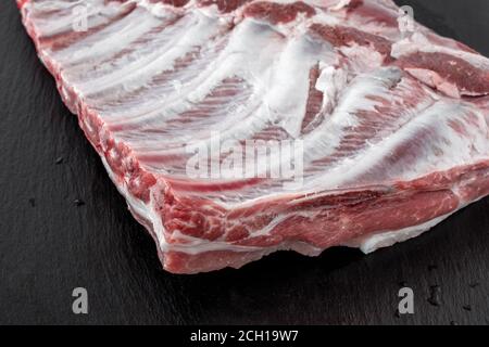 Ganze rohe Schweinefleischrippen isoliert auf dunklem, schwarzem Steinbrett. Spare Rippen oder Bauch. Nahaufnahme Metzgerei, Markt, Fleisch-Konzept Stockfoto