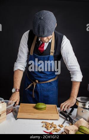 Koch in Mütze und blaue Schürze in der Küche neben dem Holztisch stehen. Auf dem Tisch auf einem Schieferteller liegt grüner appetitlich Burger Stockfoto