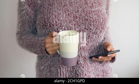 Eine Frau in einem zotteligen Pullover hält eine Tasse Kaffee oder Tee und ein Telefon in der Hand. Mädchen durchstöbern Telefon tragen Pullover mit Tasse in der Hand. Stockfoto