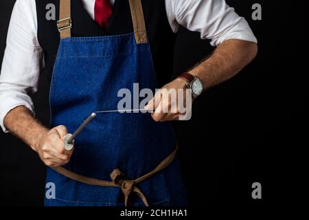 Nahaufnahme der mittleren Sektion des nicht erkennbaren Koch Koch in eleganten Uniform tragen Kappe und Schürze schärft Messer im Restaurant auf schwarzem Hintergrund. Stockfoto