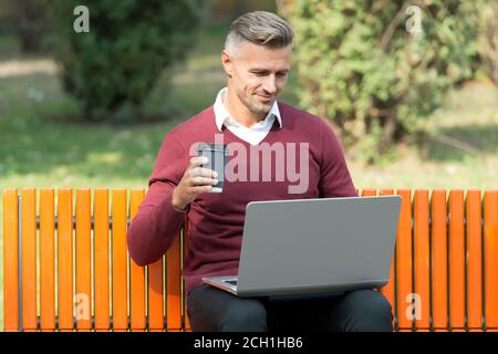 Schöner Start in den Tag. Guten Morgen. reifen schönen Mann arbeiten auf Computer im Freien. Gut gepflegten Mann sitzen auf der Bank mit Kaffee. Zuversichtlich Geschäftsmann ist online. Casual Business Fashion. Agile Business. Stockfoto