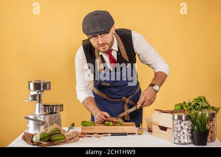 Kaukasische bärtigen Mann in Schürze und Mütze, Hamburger, Hinzufügen von Zutaten auf grüne Bun, isolierte studio Schuß auf gelbem Hintergrund Stockfoto
