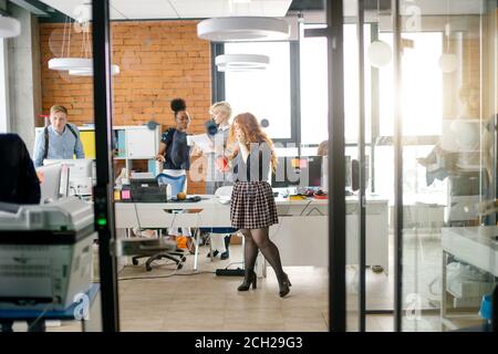 Ingwer Sekretärin in casual Clothers stehend mit Tee und Zelle Telefon in der Mitte des stark frequentierten Raumes Stockfoto