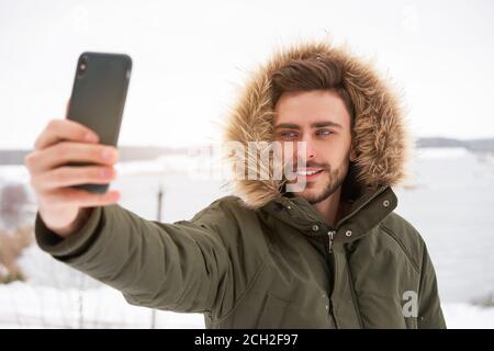 Gutaussehender bärtiger junger kaukasischer Mann steht draußen Pelze Kapuze Winter gefrorener See Glücklich lächelnd Attraktiv europäischer Kerl gehen mit selfie Stockfoto