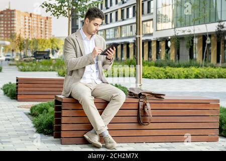Junger eleganter, erholsamer Geschäftsmann in formalwear, der auf einer Holzbank sitzt Im Park Stockfoto