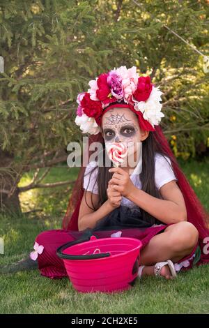 Nettes Mädchen mit Farbe auf Gesicht und große Lollypop in Hände sitzen auf grünem Gras Stockfoto