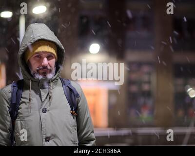 Reife kaukasischen bärtigen Mann in Winterjacke unter Schneefall stehen Und Blick auf die Kamera Stockfoto