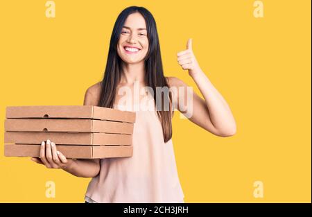 Junge schöne kaukasische Frau hält Lieferung Pizza Box lächelnd glücklich und positiv, Daumen nach oben tun ausgezeichnet und Genehmigung Zeichen Stockfoto