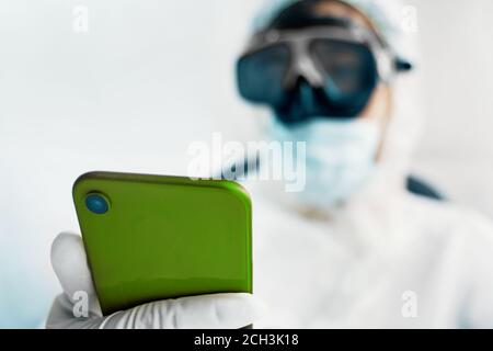 Mann in Schutzanzug Maske und Handschuhe nutzt ein grünes Smartphone für die Kommunikation mit der Familie. Epidemische Pandemie neue schnell verbreitete Coronavirus 2019-ncov, Medizin Grippe-Virus-Konzept. Stockfoto