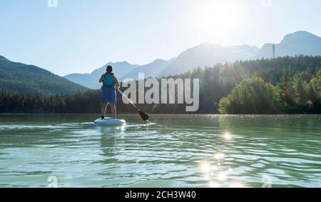 Rückansicht einer barfuß-Frau in Kleid und Schwimmweste, die an sonnigen Sommertagen in den Bergen in British Columbia, Kanada, auf einem SUP-Board auf ruhigem Seewasser reitet Stockfoto