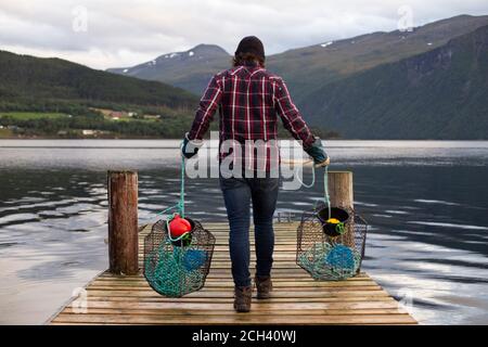 Ein Mann trägt zwei Krabbentöpfe entlang einer Anlegestelle Ein Fjord Stockfoto