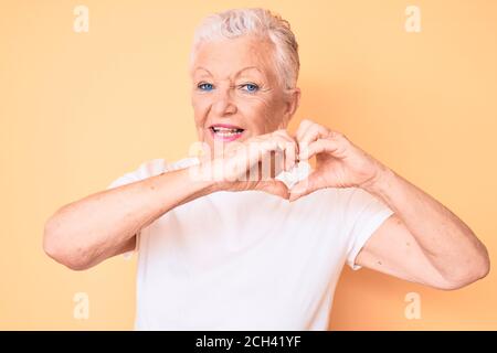 Ältere schöne Frau mit blauen Augen und grauen Haaren tragen Klassisches weißes T-Shirt auf gelbem Hintergrund lächelnd in Liebe tun Herz Symbol Form mit Stockfoto