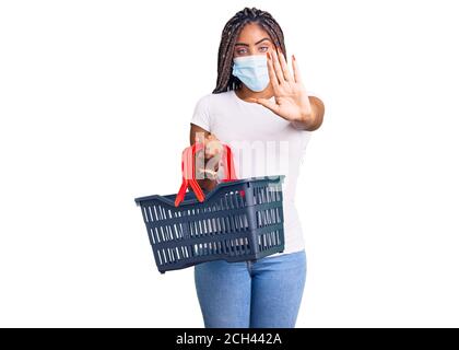 Junge afroamerikanische Frau mit Zöpfen tragen Warenkorb und Medizinische Maske mit offener Hand Stop-Zeichen mit schweren tun Und selbstbewusster Ausdruck Stockfoto