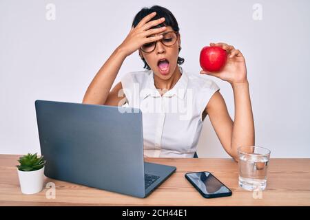 Schöne brunettte Frau arbeitet im Büro essen gesunde Apfel gestresst und frustriert mit Hand auf Kopf, überrascht und wütend Gesicht Stockfoto