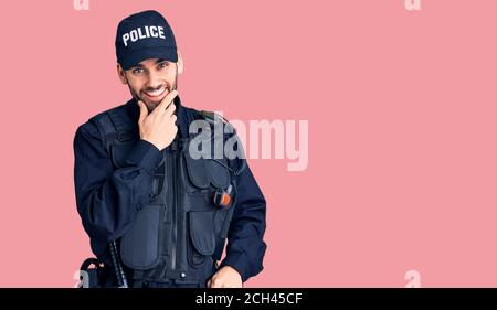 Junger gutaussehender Mann mit Bart in Polizeiuniform, der selbstbewusst auf die Kamera schaut, lächelt mit gekreuzten Armen und erhobener Hand am Kinn Stockfoto