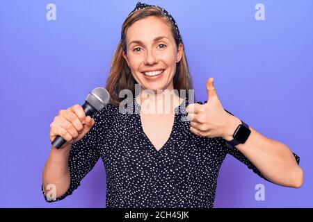 Junge schöne blonde Frau hält Mikrofon singen über lila Hintergrund lächelnd glücklich und positiv, Daumen nach oben tun ausgezeichnet und Genehmigung Zeichen Stockfoto