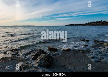 Felsen im Vordergrund mit Meereswellen, die über sie Rollen. Der blaue Ozean hat kleine Wellen über dem Wasser. Der Himmel ist blau mit weißen Wolken. Stockfoto