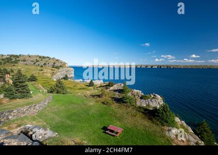 Ein abgenutzter Fußweg zu einem roten Picknicktisch auf einer Wiese an einer Küste nahe dem Rand eines blauen Ozeanwassers. Stockfoto