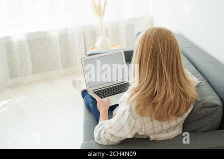 Rückansicht der unkenntlich reifen Frau mit Laptop, während sie auf einem bequemen Sofa sitzt, häusliche Innenausstattung. Schöne Frauen einkaufen oder chatten online in sozialen Netzwerk, freiberuflich arbeiten Stockfoto