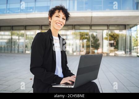 Porträt von lächelnden reifen Geschäftsfrau in Anzug mit Laptop, während Sitzen außerhalb des Büros Stockfoto