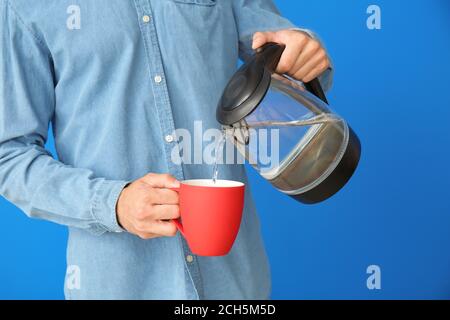Junger Mann gießt Wasser aus dem Wasserkocher in die Tasse Farbiger Hintergrund Stockfoto