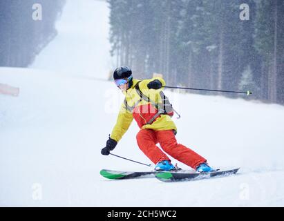 Vorderansicht des jungen männlichen Skifahrers Abfahrtsski und tun Carve Turn auf hohen schneebedeckten Hang. Skifahren bei Schneefall in den Bergen. Einfache Abfahrt. Winter nebligen Hintergrund. Aktives sportliches Lifestyle-Konzept. Stockfoto