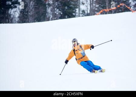 Der professionelle Skifahrer konzentrierte sich auf das Skifahren auf der steilen Skipiste. Geübtes technisches Carving Skifahren. Netz an Hangkante. Bewaldete Bergspitze im Hintergrund. Extreme Winteraktivitäten Konzept Stockfoto