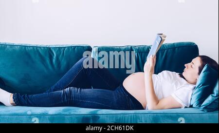 Startseite Gemütliches Porträt der schwangeren Frau zu Hause ruhen und Buch lesen. Schwanger mittleren Alters Mutter Porträt liegend auf Sofa zu Hause. Gesunde Schwangerschaft c Stockfoto