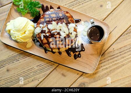 Draufsicht auf Bananenpfannkuchen mit Macadamianuss und Schokoladensirup auf Pfannkuchen-Stapel und Vanilleeis auf Holzplatte auf Holztisch. Stockfoto