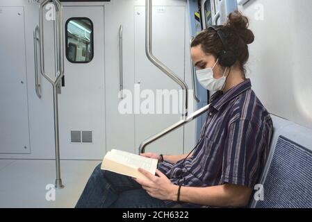 Isoliert und maskiert smart aussehende junge Erwachsene Mann hört zu Musik und Lesen eines Literaturbuches während der Reise in der Blick auf die U-Bahn-Seite Stockfoto