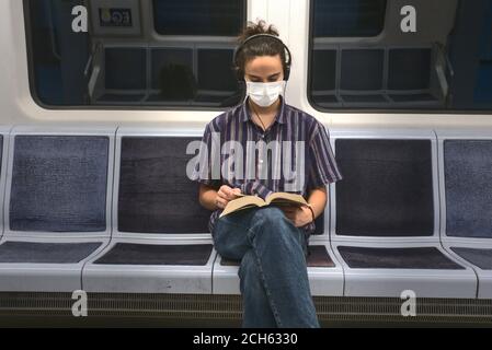 Isoliert und maskiert smart aussehende junge Erwachsene Mann hört zu Musik und Lesen eines Literaturbuches während der Reise in der U-Bahn Stockfoto