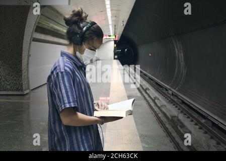 Isoliert maskiert langhaarige junge Erwachsene Musik aus hören Sein Kopfhörer und Literatur Buch lesen, während auf die warten Die U-Bahn kommt Stockfoto