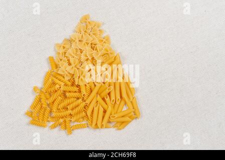 Verschiedene rohe Pasta-Typen in Form eines Dreiecks auf rustikalem Leinenstoff ausgelegt, Draufsicht. Verschiedene Formen von Pasta fusilli, Penne und farfalle Stockfoto