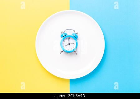 Blauer Wecker und leerer Teller auf farbigem Papierhintergrund. Intermittierendes Fasten Konzept - Bild Stockfoto